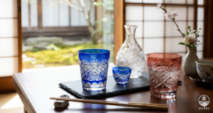 A serene indoor scene featuring handcrafted blue and red Kiriko (Japanese cut glass) sake glasses and a decanter on a wooden table. Natural daylight from a shoji screen window and a traditional Japanese garden background highlight the intricate geometric patterns. Chopsticks and a floral arrangement complete the refined table setting.