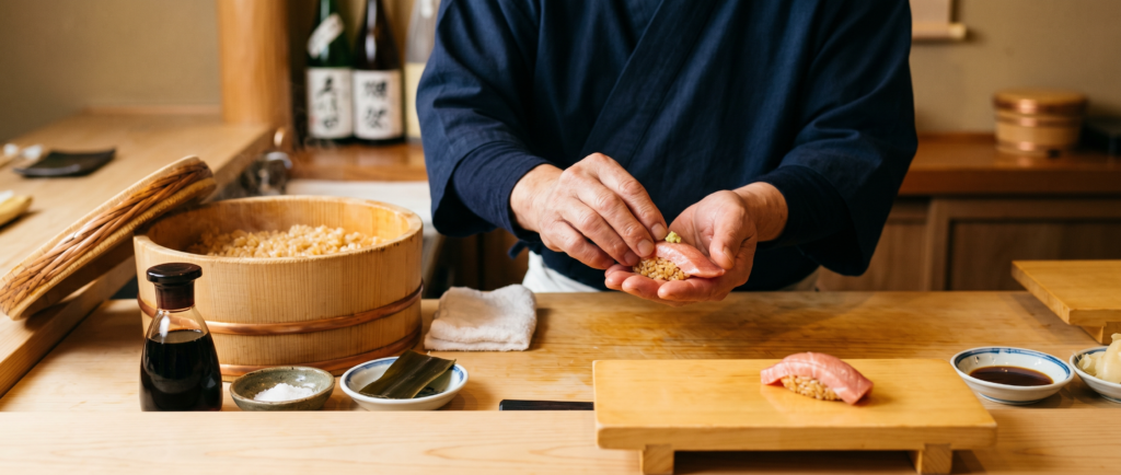 Sushi chef shaping nigiri by hand at a counter, with a wooden hangiri tub of seasoned sushi rice (shari) in the foreground