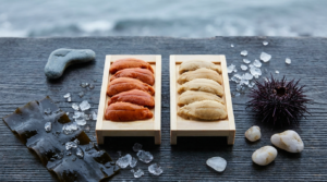 Two wooden trays of sea urchin roe, one deep orange and one pale cream-gold, beside fresh konbu kelp on a dark weathered cedar board.